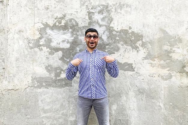 Portrait of surprised handsome bearded young man in checkered blue shirt and sunglasses standing against concrete gray wall, pointing himself, asking and looking with shocked amazed face.