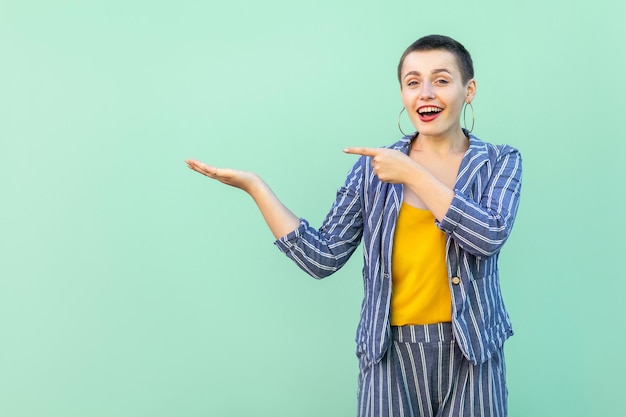 Portrait of surprised handsome beautiful short hair young stylish woman in casual striped suit standing amazed, pointing at something on hand. indoor studio shot isolated on light green background.