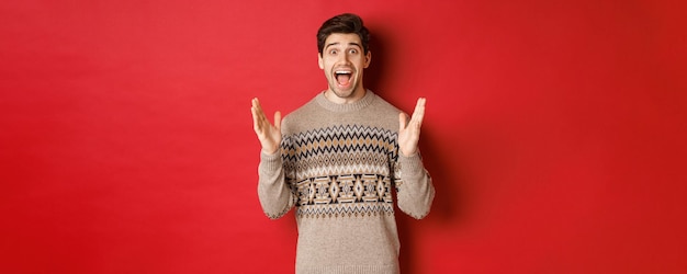 Portrait of surprised and happy handsome man, wearing christmas sweater, looking amazed, celebrating new year, standing over red background