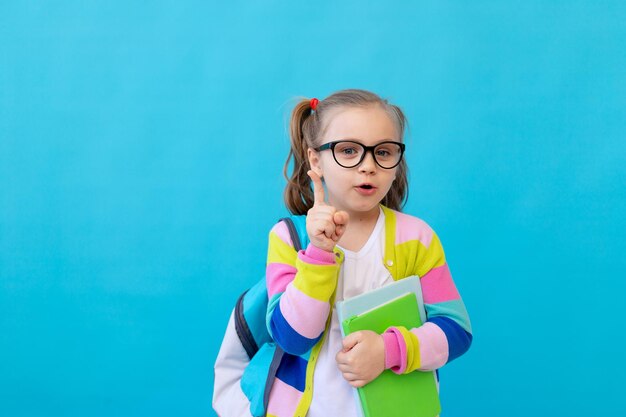 Portrait of a surprised little girl with glasses in a striped jacket with notebooks and textbooks in her hands and a backpack. The concept of education. Photo studio, blue background, place for text.