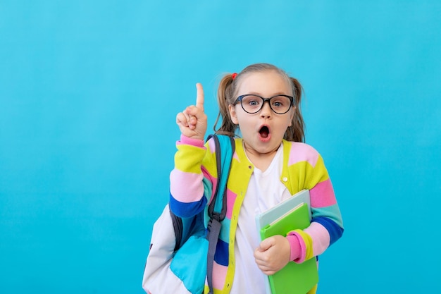 Портрет удивленной маленькой девочки в очках в полосатой куртке с тетрадями и учебниками в руках и рюкзаком Концепция образования Фотостудия синий фон место для текста