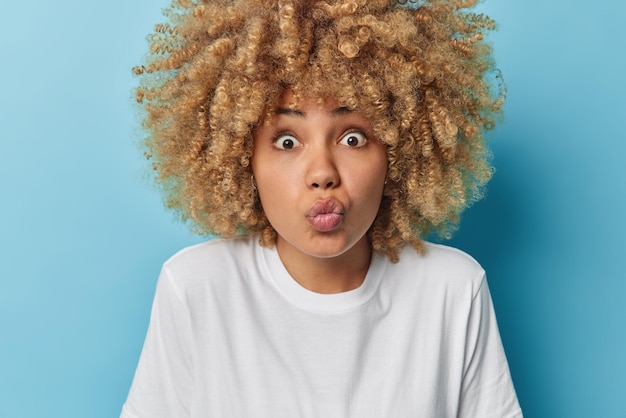 Portrait of surprised lovely woman has widen eye lips folded focused at camera feels shocked dressed in casual white t shirt stands against blue background People and facial expressions concept