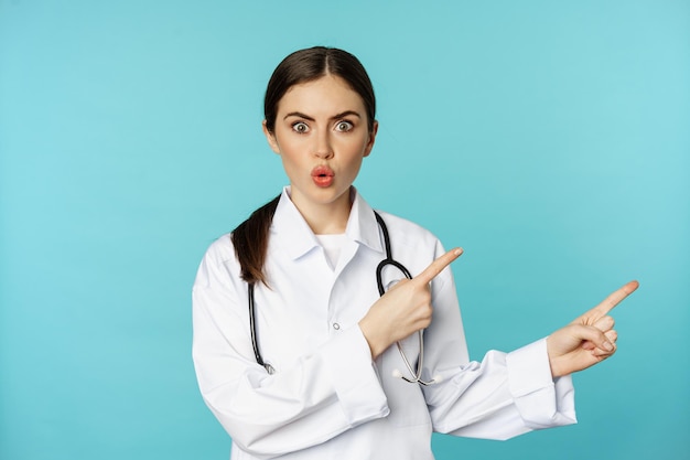 Portrait of surprised medical worker, woman doctor pointing fingers right and say wow amazed, reacting interested, standing over torquoise background