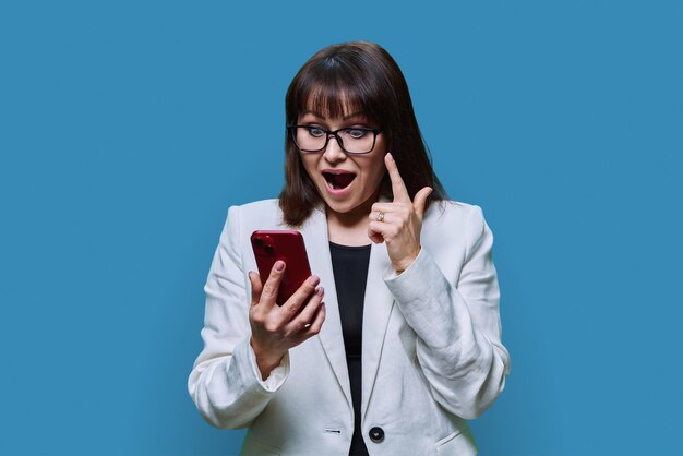 Photo portrait of surprised middle aged business woman with phone on blue background