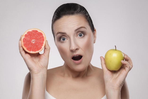 Portrait of surprised middle aged woman with perfect face skin holding fresh slice grapefruit and apple in her hands and open mouth, isolated on grey background. Indoor, studio shot, copy space
