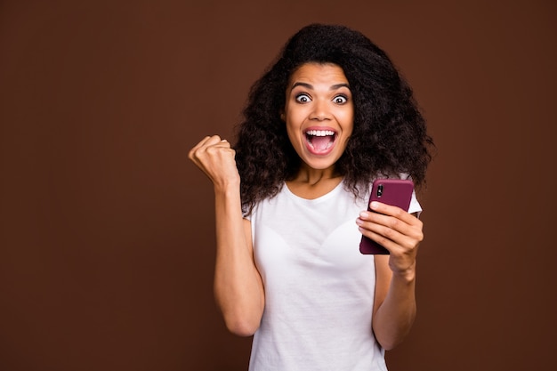 Portrait of surprised shocked afro american girl use smart phone read win lottery social network  news impressed scream wow omg wear white t-shirt.