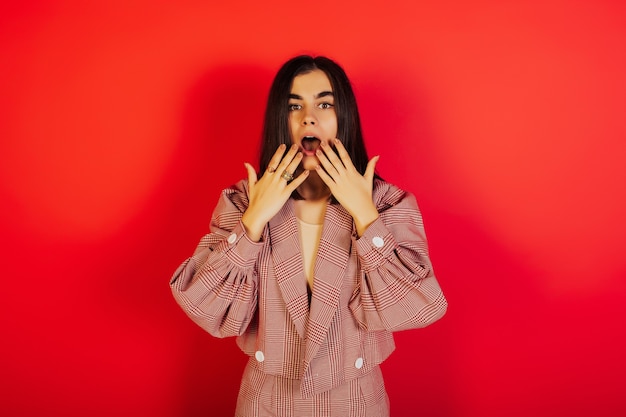 Portrait of surprised and stunned stylish woman in red clothes, holding hands near mouth while staring at camera over red surface.