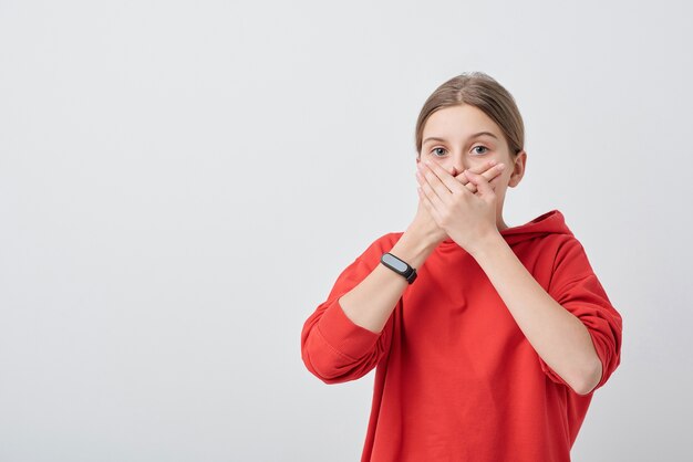 Portrait of surprised teenaged girl in hoodie covering mouth with hands in excitement