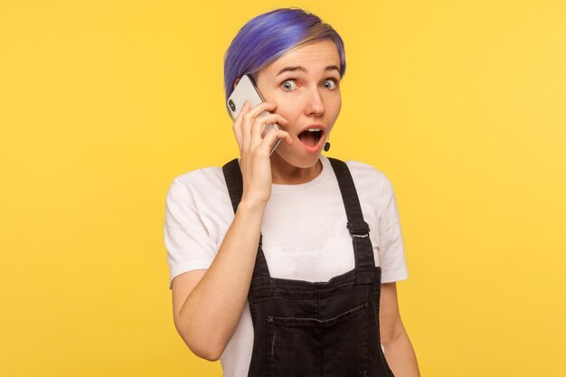 Portrait of surprised trendy hipster girl with violet short hair in overalls calling and talking on cell phone shocked after hearing unbelievable news looking amazed yellow background studio shot