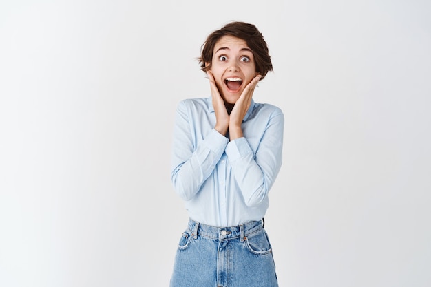 Portrait of surprised woman scream happy and joyful, checking out amazing promo offer, gasping and holding hands near opened mouth, standing on white wall