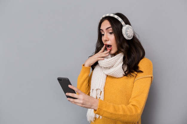 Portrait of surprised woman wearing ear muffs and scarf holding mobile phone, isolated over gray background