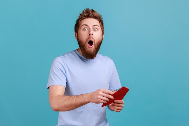 Portrait of surprised young adult handsome bearded man reading letter or greeting card holding envelope being astonished of shocked news Indoor studio shot isolated on blue background