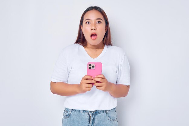 Портрет удивленной молодой азиатки в белой футболке с помощью мобильного телефона на белом фоне