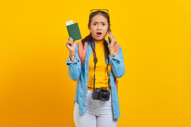 Portrait of surprised young Asian traveler holding boarding pass passport ticket and talking on smartphone isolated on yellow background. Passengers traveling on weekends. Air flight travel concept