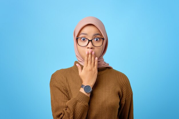 Portrait of surprised young asian woman covering mouth with hand on blue background