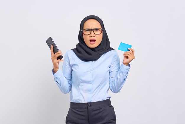 Portrait of surprised young Asian woman holding mobile phone and showing credit card isolated on white background