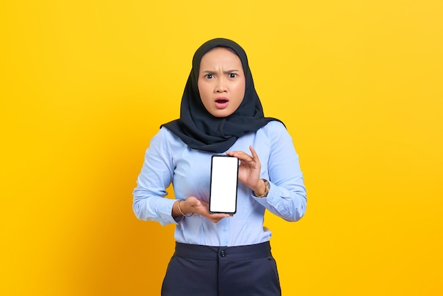黄色の背景で隔離の空白の画面の携帯電話を示す驚いた若いアジアの女性の肖像画