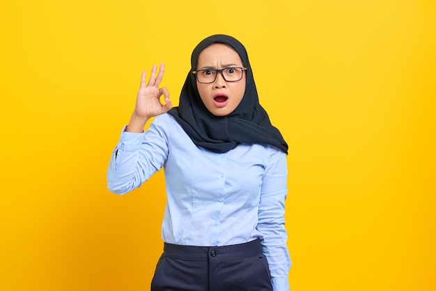 Portrait of surprised young Asian woman showing okay gesture isolated on yellow background