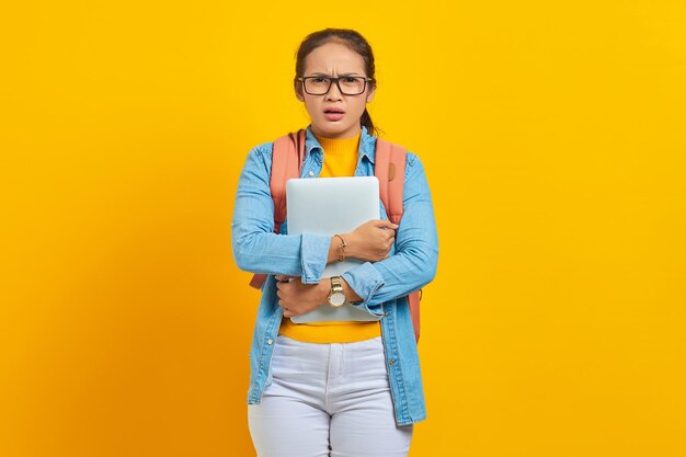 Portrait of surprised young asian woman student in casual clothes with backpack holding laptop and looking at camera isolated on yellow background education in university college concept