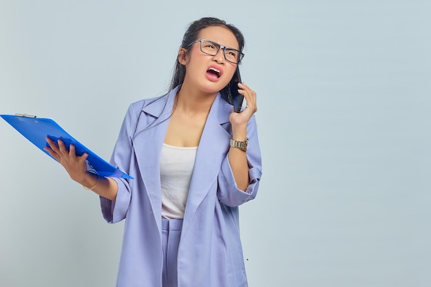 Портрет удивленной молодой азиатки, разговаривающей по мобильному телефону и просматривающей список задач в папке документов, изолированной на фиолетовом фоне