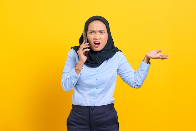 Portrait of surprised young Asian woman talking on mobile phone with open mouth isolated on yellow background