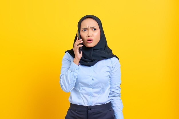 Portrait of surprised young asian woman talking on mobile phone\
with open mouth isolated on yellow background