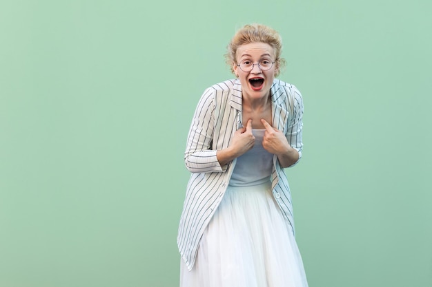 Portrait of surprised young blonde woman in white striped blouse with eyeglasses standing, amazed face, pointing herself and looking at camera. studio shot isolated on light green background.
