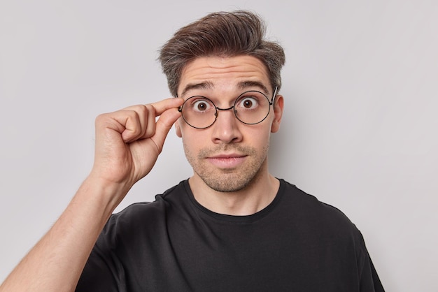 Portrait of surprised young man keeps hand on rim of spectacles stares amazed hears unbelievable news dressed in casual black t shirt isolated over white background. Human reactions concept.