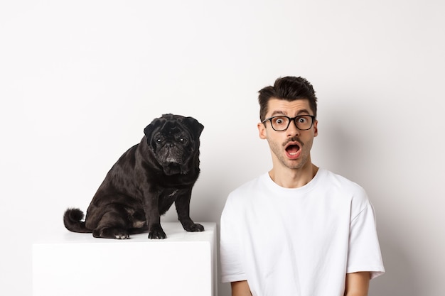 Portrait of surprised young man staring at camera, sitting near his dog pug, posing over white background.
