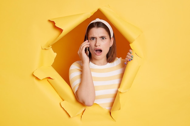 Portrait of surprised young woman wearing striped tshirt and\
hair band looking at camera with big eyes and talking on cell phone\
breaks through yellow paper background in ripped hole