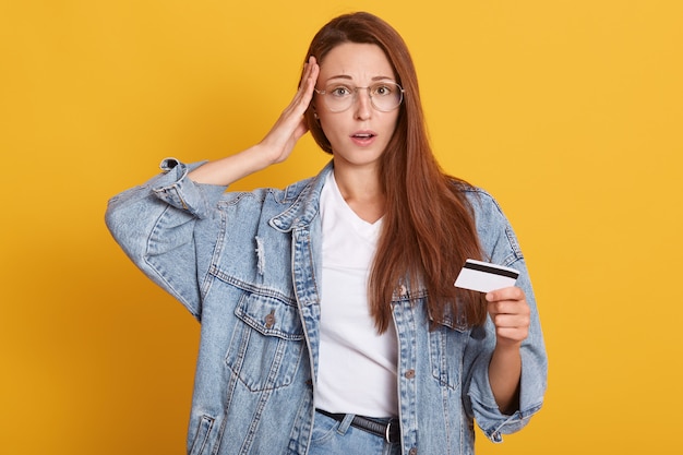 Portrait of surprised young woman wears casual clothes keeps mouth open and putting palm on temple, holding credit bank card in hand