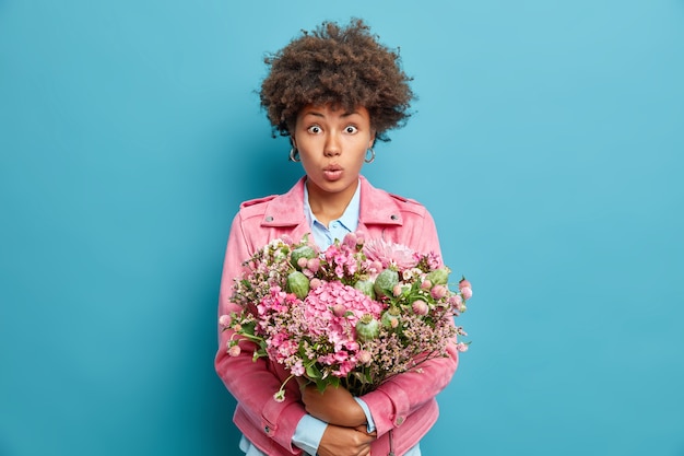 Portrait of surprised young woman with curly hair embraces big nice bunch of flowers shocked to get congratulations on birthday isolated over blue wall