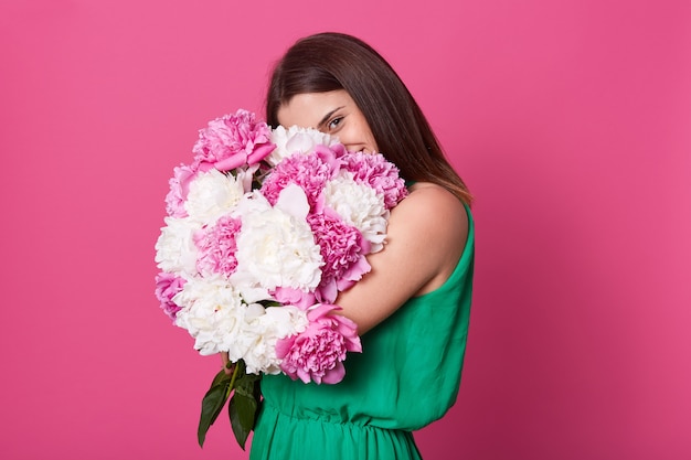 Portrait of sweet attractive female holding huge bouquet of peonies close to her