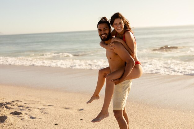 Portrait of a tanned Caucasian couple on holiday, having fun piggybacking on a sunny beach, looking to camera and smiling, with blue sky and sea in the background