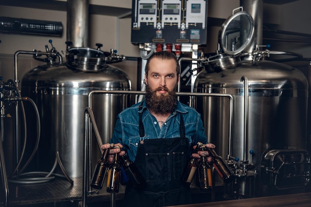 Portrait of tattooed, bearded hipster male manufacturer presenting beer in the microbrewery.