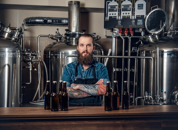 Photo portrait of tattooed, bearded hipster male manufacturer presenting beer in the microbrewery.