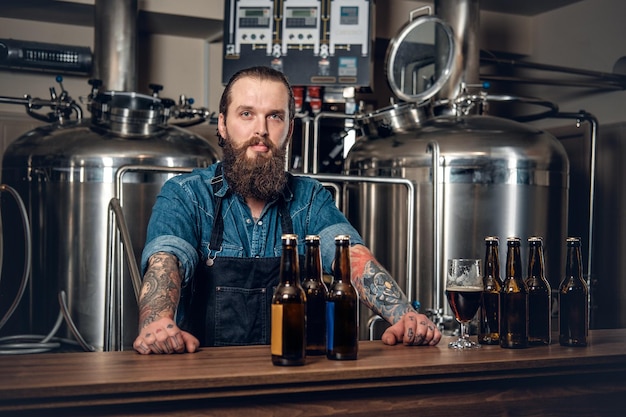 Portrait of tattooed, bearded hipster male manufacturer presenting beer in the microbrewery.