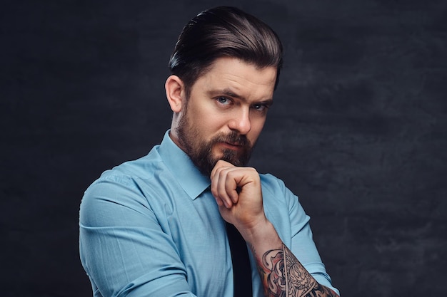 Portrait of a tattooed handsome middle-aged man with beard and hairstyle dressed in a blue shirt and tie, pose in a studio with crossed arms. Isolated on a textured dark background in studio.