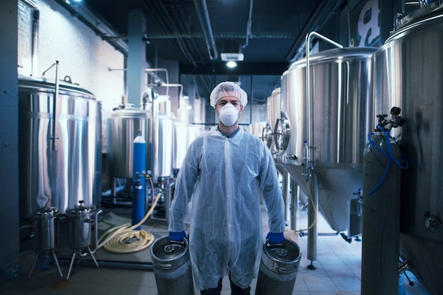 Portrait of technologist in white uniform with hairnet and mask holding gas bottles in plant for food or beverages production.