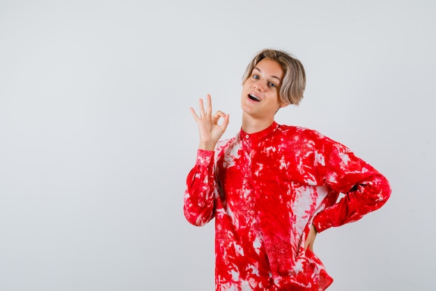 Portrait of teen blonde male showing ok gesture in oversized shirt and looking merry front view
