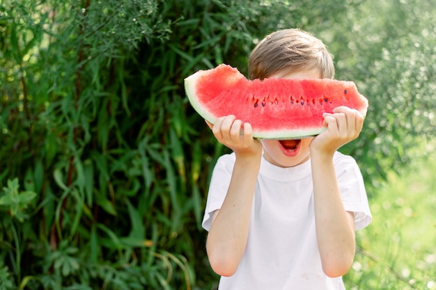 熟したジューシーなスイカを食べて笑っている十代の少年の肖像画。かわいい白人の若いティーンエイジャー。面白い幸せな子供はスライス赤いスイカで彼の顔を覆った。