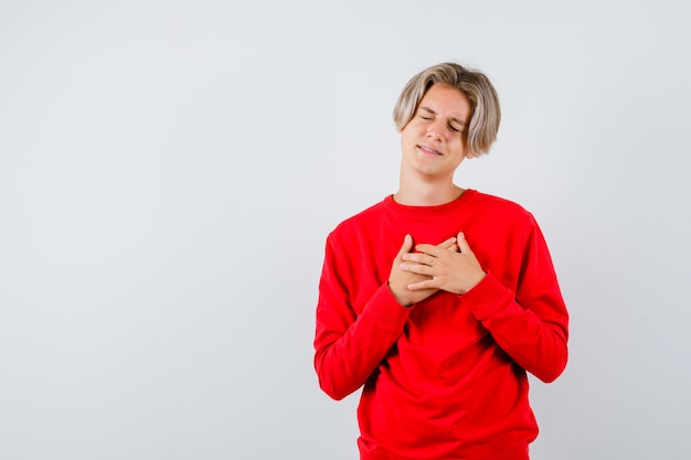 Portrait of teen boy with hands on chest while closing eyes in red sweater and looking sorrowful front view