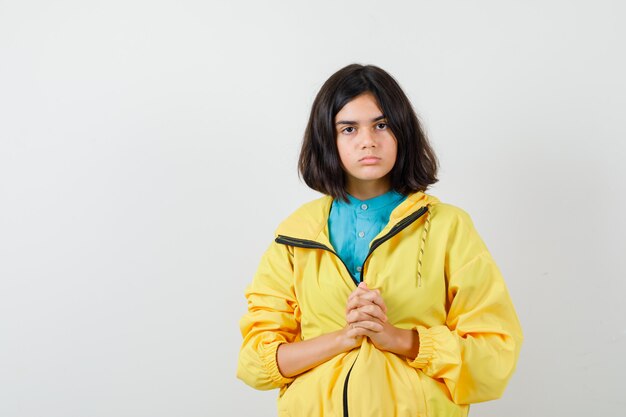 Portrait of teen girl holding fingers intertwined in yellow jacket and looking puzzled front view