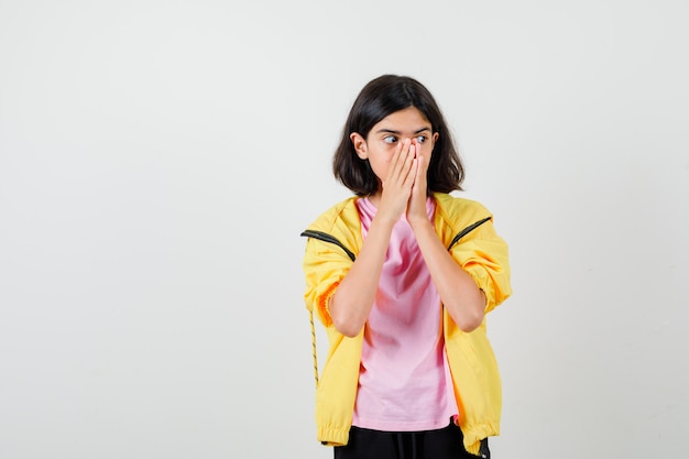 Portrait of teen girl keeping hands on face in t-shirt, jacket and looking scared front view