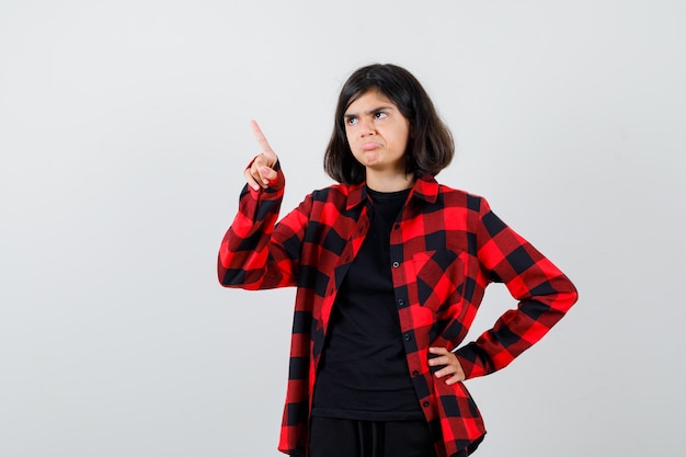 Ritratto di una ragazza adolescente che punta lontano, tenendo la mano sulla vita in una camicia casual e guardando la vista frontale sconvolta