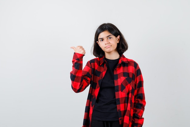 Portrait of teen girl pointing left with thumb in casual shirt and looking upset front view