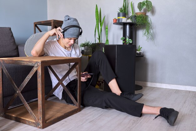 Portrait of teenage boy 16 years old in headphones listening to music, audiobooks, lessons. Home interior indoor plants cacti potted background