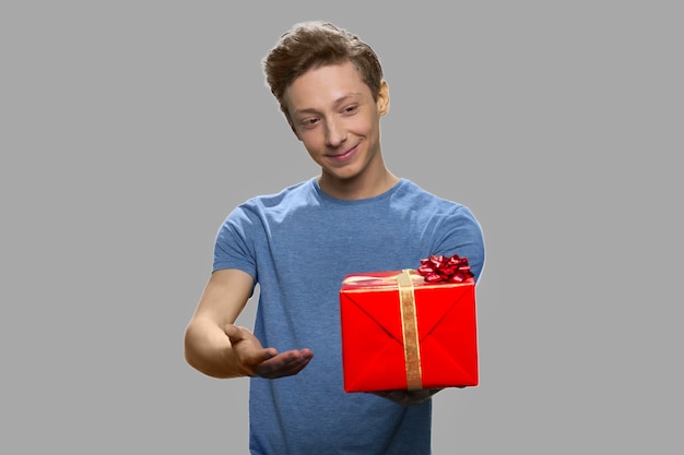 Portrait of teenage boy holding gift box. Cute boy in blue t-shirt offering present box on gray background. Special holiday offer.