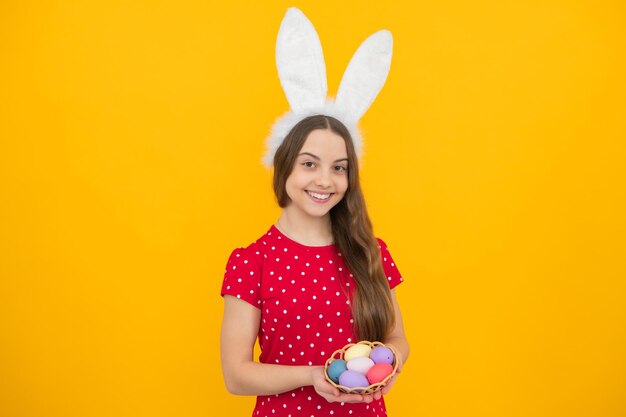 Portrait of teenage girl child wearing bunny ears holding easter eggs isolated at yellow background