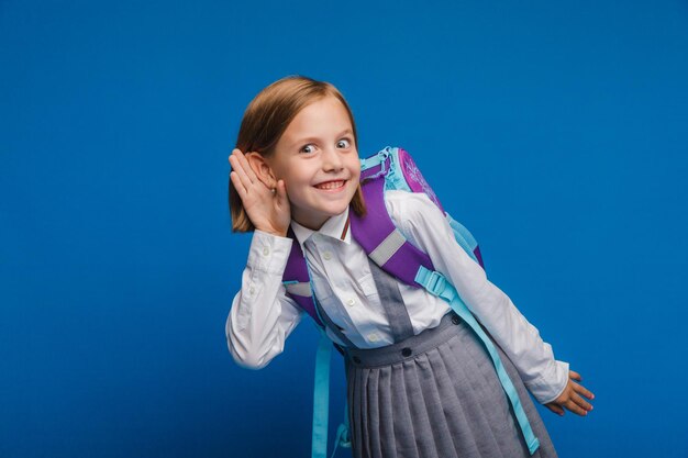 Портрет девочки-подростка, держащей руку у уха и слушающей что-то изолированное на синем фоне, милая школьница подслушивает